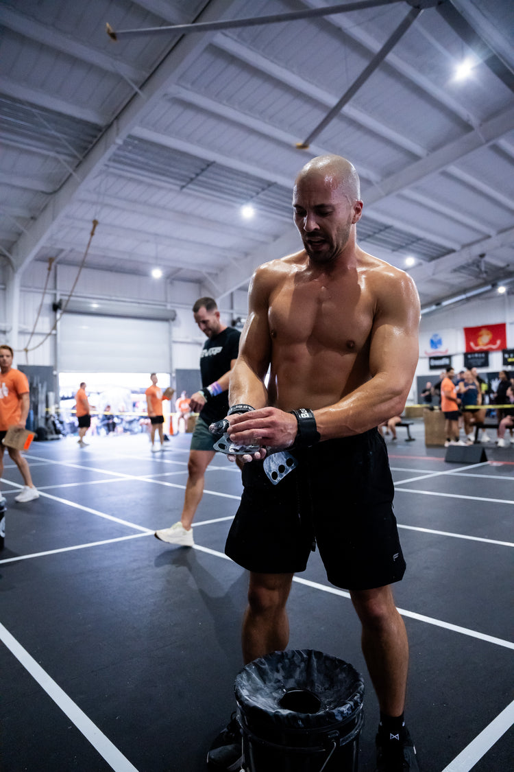 Athlete preparing for a CrossFit competition workout, standing shirtless on a gym floor while adjusting his hand grips above a chalk bucket. The image captures the focus, determination, and intensity of competitive functional fitness.