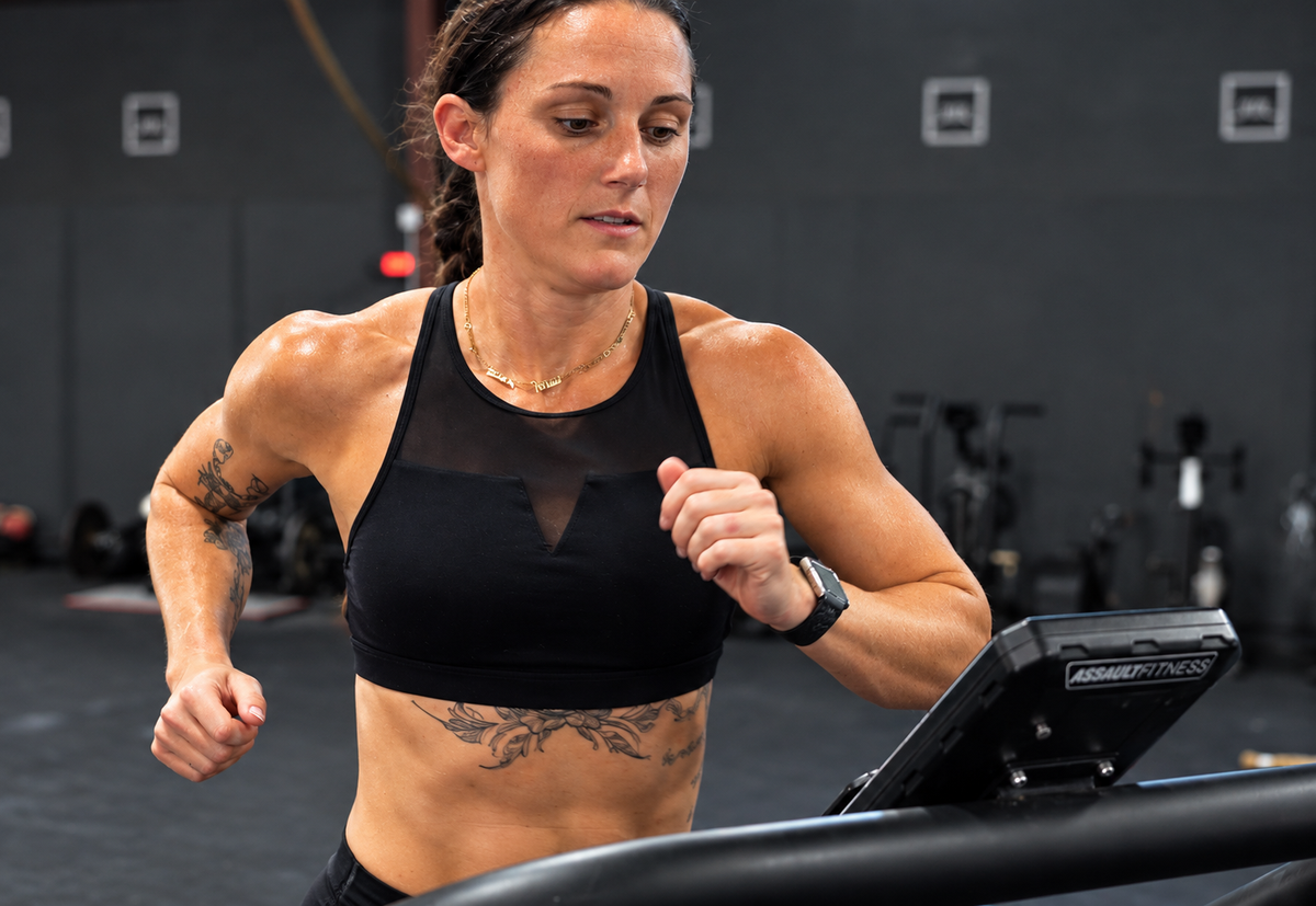 Athletic woman with tattoos running on an Assault Fitness treadmill in a gym, wearing black workout apparel and a smartwatch while maintaining a focused expression