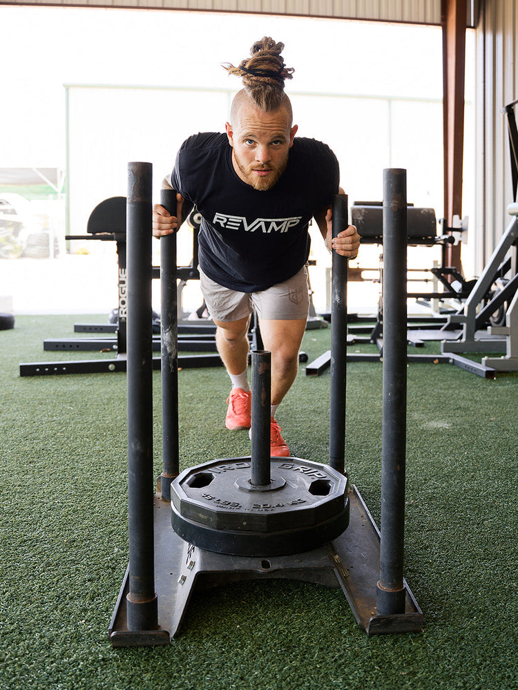 A man in a gym wearing a black REVAMP t-shirt and gray shorts, pushing a heavy black sled with a weight plate on green turf